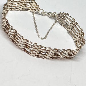 Puede incluir: Una pulsera plateada con un diseño tejido. La pulsera presenta una serie de eslabones rectangulares interconectados, creando una superficie texturizada. Una pequeña cadena está unida al cierre para mayor seguridad. La pulsera se muestra sobre una superficie blanca.