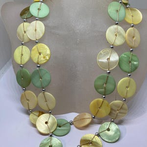 Könnte beinhalten: Eine zweireihige Halskette aus großen, runden, irisierenden Muschelknäpfen in Grün, Gelb und Creme. Die Knäpfe sind auf einer dünnen, goldenen Schnur mit silbernen Perlen zwischen jedem Knopf aufgefädelt.