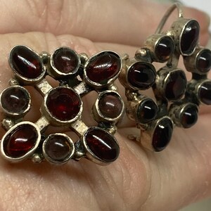 May include: A pair of vintage silver earrings with dark red, oval-shaped gemstones. One earring is a square shape with gemstones set in a grid pattern. The other earring is a cluster of gemstones. The earrings are displayed against a neutral background.
