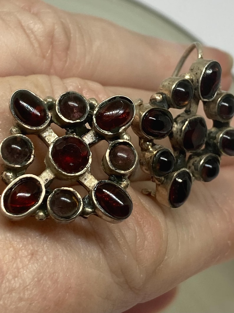 May include: A pair of vintage silver earrings with dark red, oval-shaped gemstones. One earring is a square shape with gemstones set in a grid pattern. The other earring is a cluster of gemstones. The earrings are displayed against a neutral background.