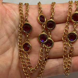 May include: Gold-tone necklace featuring a series of round, deep purple gemstones set in gold bezels. The necklace has a multi-strand chain design, with the gemstones evenly spaced along the chains. The chain is a classic link style.