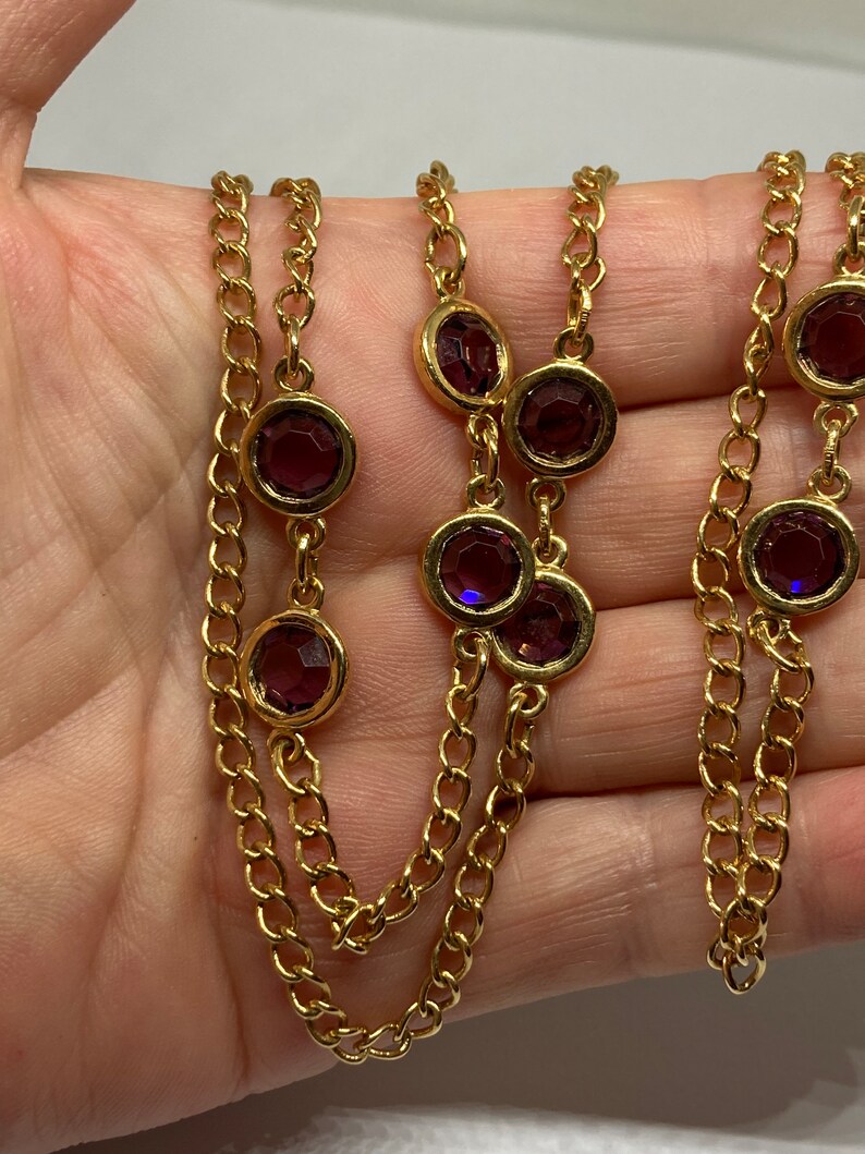 May include: Gold-tone necklace featuring a series of round, deep purple gemstones set in gold bezels. The necklace has a multi-strand chain design, with the gemstones evenly spaced along the chains. The chain is a classic link style.