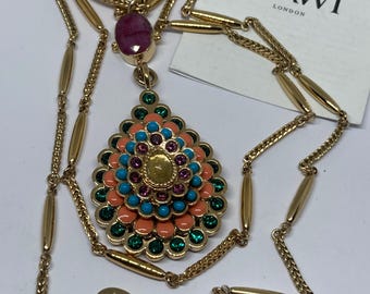 Puede incluir: Un collar de oro con un colgante multicolor con un diseño rosa, turquesa y verde. El colgante está rodeado por un círculo de piedras verdes. El collar tiene un cierre de langosta y una etiqueta que dice "MAWI LONDON".