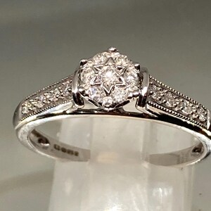May include: A silver ring with a starburst diamond cluster center and smaller diamonds along the band. The ring is set on a clear surface, showcasing its intricate design and sparkle. A classic piece of jewelry.