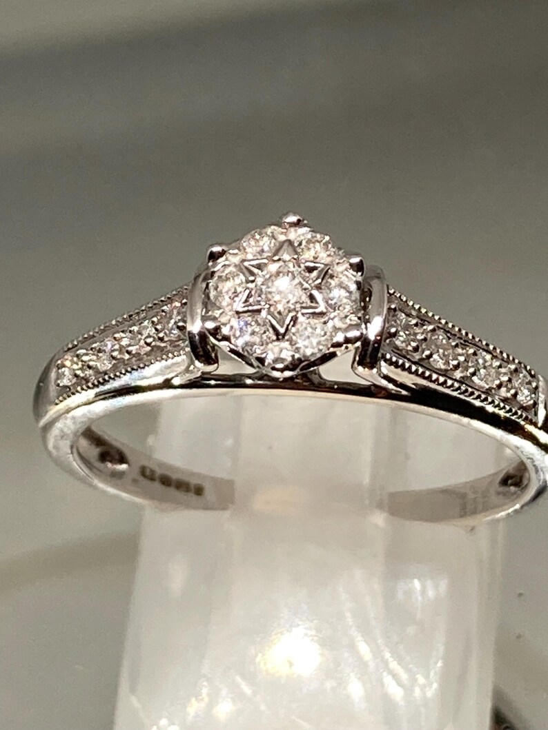 May include: A silver ring with a starburst diamond cluster center and smaller diamonds along the band. The ring is set on a clear surface, showcasing its intricate design and sparkle. A classic piece of jewelry.