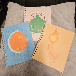 May include: Three spiral-bound notebooks with colorful illustrations. One has an orange and yellow design, another features a green and pink design, and the third has an orange design on a blue background. The notebooks are on a gray surface.