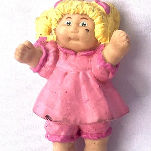 Puede incluir: Una pequeña figura de plástico de una muñeca Cabbage Patch Kid. La muñeca lleva un vestido rosa y tiene el pelo rubio con lazos rosas.