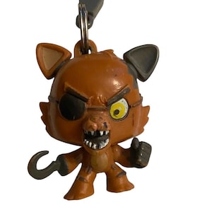 Könnte beinhalten: Ein brauner Foxy the Pirate Schlüsselanhänger mit Hakenhand, Augenklappe und bedrohlichem Gesichtsausdruck. Der Schlüsselanhänger hat einen grauen Verschluss und Ring. Die Figur stammt aus dem Franchise Five Nights at Freddy's.