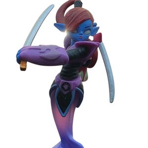 May include: A figurine of a blue-skinned character with red hair, pointy ears, and two swords. The figure is wearing a purple and black outfit and stands on a patterned base with an orange border. The character is in a dynamic pose.