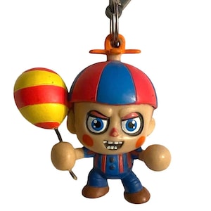 Könnte beinhalten: Ein Schlüsselanhänger mit einer Cartoonfigur mit roter und blauer Mütze, blauen Overalls und einem gelb-rot gestreiften Ballon. Die Figur hat einen wütenden Gesichtsausdruck und hält den Ballon. Der Schlüsselanhänger hat einen grauen Clip.