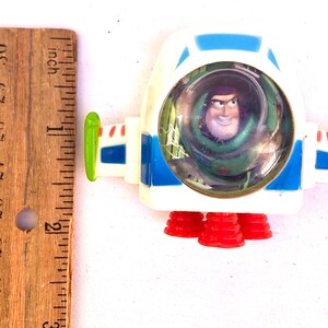 Op de afbeelding: Een wit en blauw speelgoed ruimteschip met Buzz Lightyear uit Toy Story. Het speelgoed heeft rode raketboosters en een doorzichtige cockpit. Het ruimteschip is ongeveer 5 cm lang.