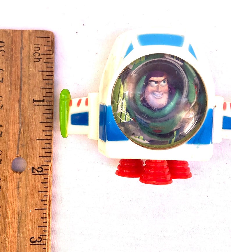 Op de afbeelding: Een wit en blauw speelgoed ruimteschip met Buzz Lightyear uit Toy Story. Het speelgoed heeft rode raketboosters en een doorzichtige cockpit. Het ruimteschip is ongeveer 5 cm lang.