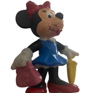 May include: A vintage Minnie Mouse figurine. The figure is black, white, and tan with a red bow, dress, shoes, and purse. She is holding a yellow object. The figurine is standing on a white surface.