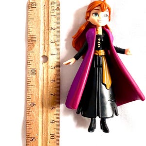 May include: A small plastic doll of Anna from the movie Frozen. She is wearing a black dress with a gold belt, a purple cape, and has long red hair. The doll is approximately 12.7 cm tall.