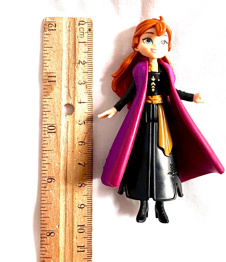 May include: A small plastic doll of Anna from the movie Frozen. She is wearing a black dress with a gold belt, a purple cape, and has long red hair. The doll is approximately 12.7 cm tall.