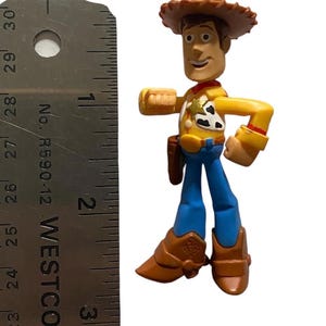 May include: A Woody action figure from the Toy Story franchise. The figure is wearing a brown cowboy hat, yellow shirt, blue pants, and brown boots. The figure is approximately 3 inches tall.