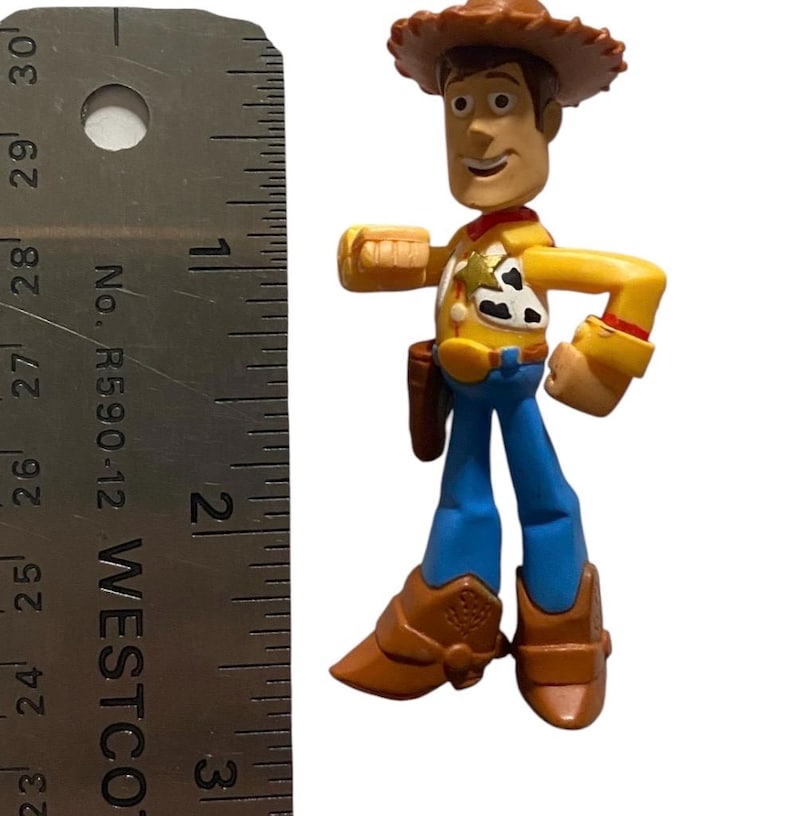 May include: A Woody action figure from the Toy Story franchise. The figure is wearing a brown cowboy hat, yellow shirt, blue pants, and brown boots. The figure is approximately 3 inches tall.