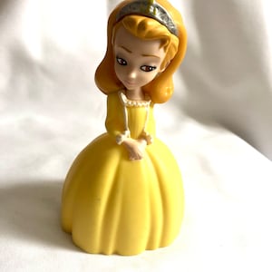 May include: A yellow plastic doll wearing a yellow dress and a silver crown. The doll has brown eyes and blonde hair.