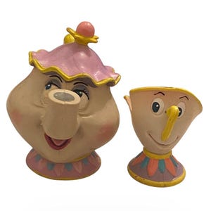 May include: A whimsical, off-white teapot and teacup set. The teapot has a pink lid with a yellow trim and a smiling face. The teacup, with a yellow handle, also has a smiling face. Both pieces have colorful bases, creating a charming, cartoonish aesthetic.