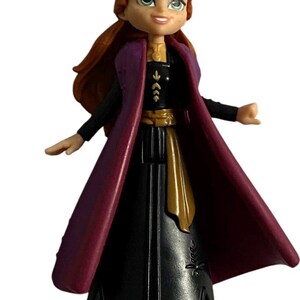 May include: A small plastic doll of Anna from the film Frozen. She is wearing a black dress with a gold belt, a purple cape, and brown boots. Her hair is red and she has blue eyes.