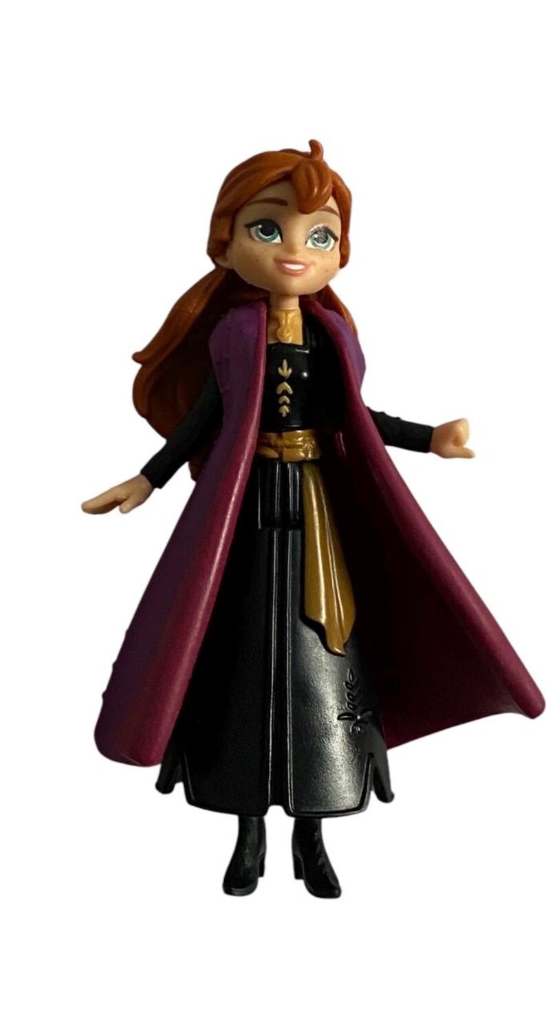 May include: A small plastic doll of Anna from the film Frozen. She is wearing a black dress with a gold belt, a purple cape, and brown boots. Her hair is red and she has blue eyes.
