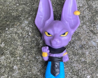 Peut inclure: Une figurine humanoïde violette ressemblant à un chat avec de grandes oreilles pointues, des yeux jaunes et une boucle d'oreille dorée. Le personnage porte un collier noir, des brassards dorés, une ceinture noire sur une jupe bleue et des bottes noires.