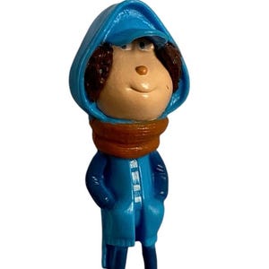 May include: A blue figurine of a person wearing a blue hooded jacket, an orange scarf, and red shoes. The figurine is standing on a red base.