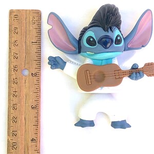 May include: Blue Stitch character figurine playing a brown guitar. Stitch is wearing a white jumpsuit with a black pompadour hairstyle. The figurine is approximately 4 inches tall.