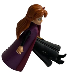 May include: A purple and burgundy dress with a black bodice and long brown hair. The doll is sitting with its legs crossed and is wearing black shoes.