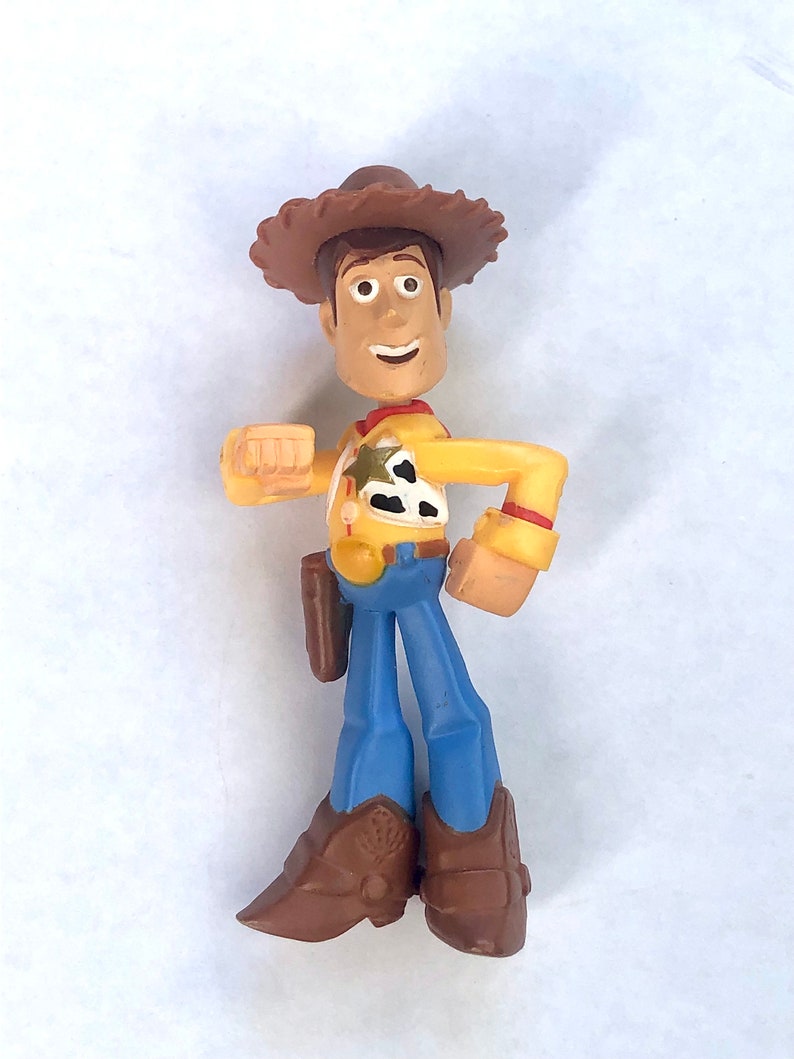 May include: A Woody action figure from the Toy Story franchise. The figure is wearing a brown cowboy hat, yellow shirt, blue pants, and brown boots. The figure has a holster and a sheriff's badge. The figure is standing on a white surface.