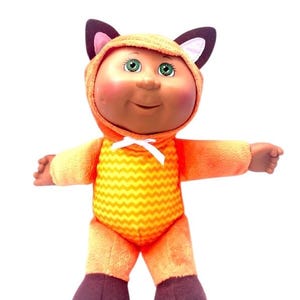 May include: A plush toy doll dressed in an orange and yellow fox costume. The doll has a brown face, green eyes, and a white bow tie.
