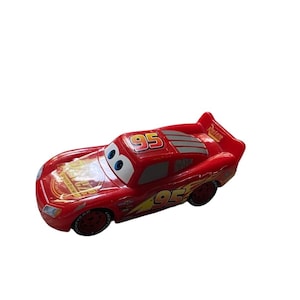May include: A red toy car featuring the number "95" on the roof and sides. The car has large, cartoonish eyes and a rear spoiler. The Lightning McQueen car is a popular character from the "Cars" film.