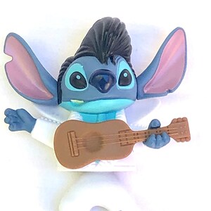 May include: Blue and white plastic figurine of Stitch from Lilo & Stitch, wearing a white jumpsuit and a black pompadour hairstyle. He is holding a brown guitar.