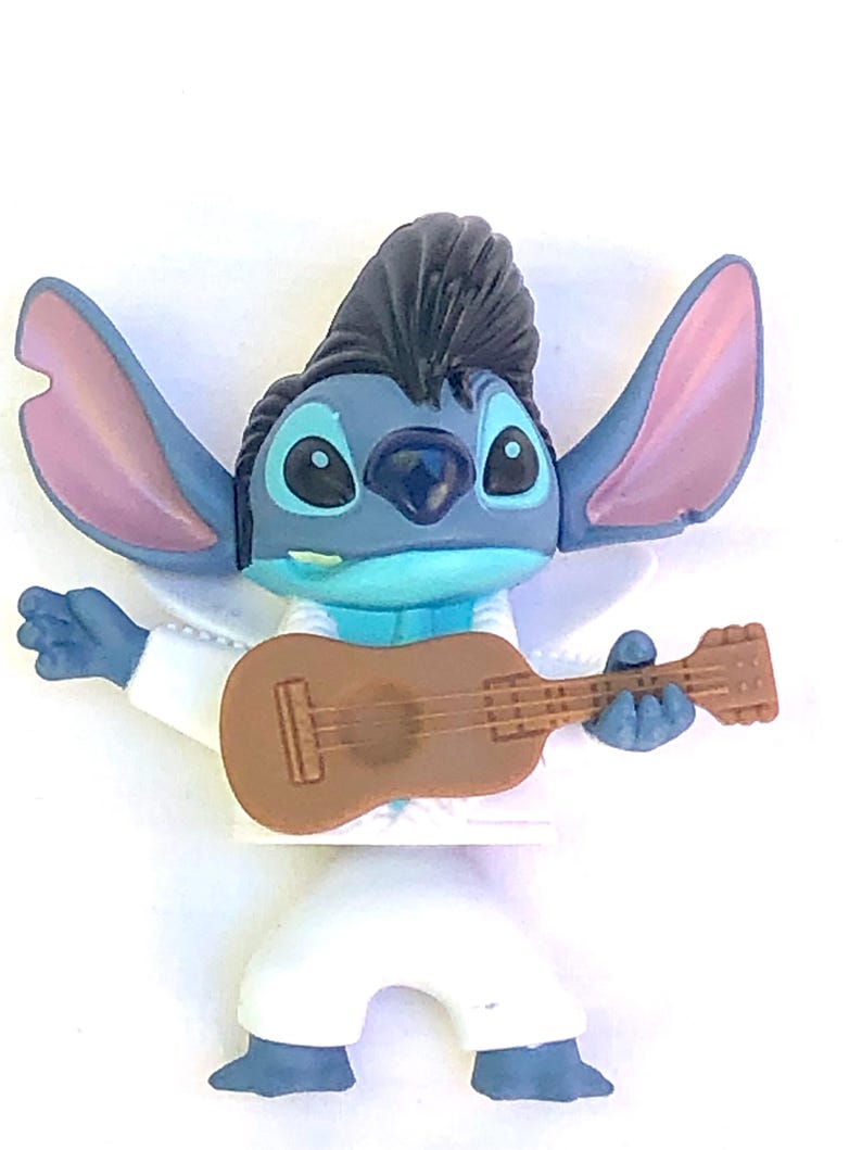 May include: Blue and white plastic figurine of Stitch from Lilo & Stitch, wearing a white jumpsuit and a black pompadour hairstyle. He is holding a brown guitar.