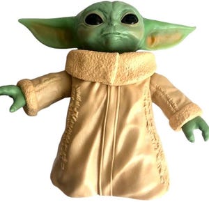 May include: A green and brown plush toy of a character from the Star Wars franchise, wearing a tan coat with a furry collar. The toy has large eyes and a serious expression.