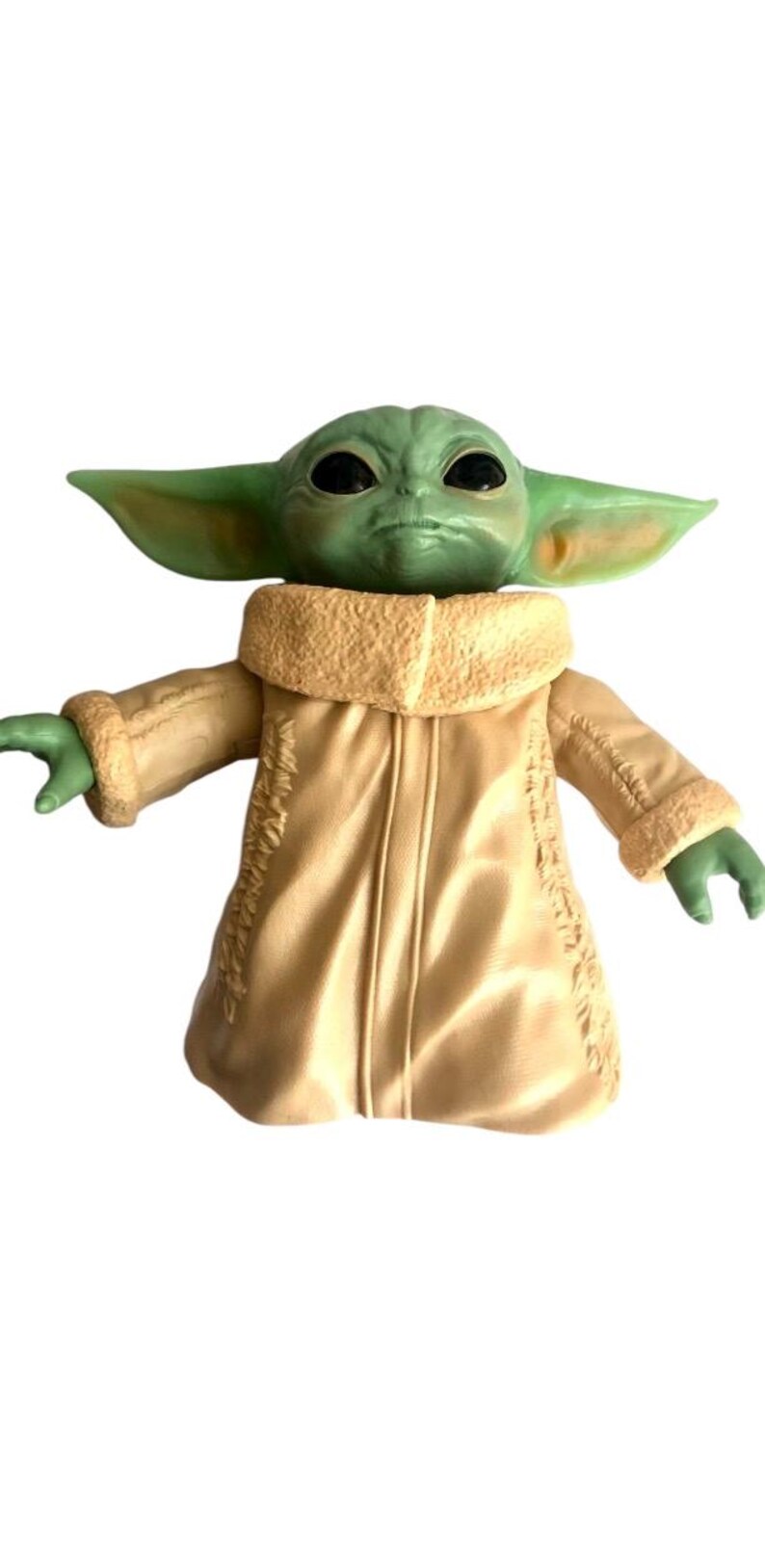 May include: A green and brown plush toy of a character from the Star Wars franchise, wearing a tan coat with a furry collar. The toy has large eyes and a serious expression.