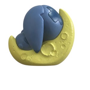 K&ouml;nnte beinhalten: Eine blaue Cartoonfigur liegt auf einem gelben Halbmond. Die Figur hat gro&szlig;e Ohren und einen runden K&ouml;rper. Der Mond hat eine strukturierte Oberfl&auml;che mit mehreren Kratern.