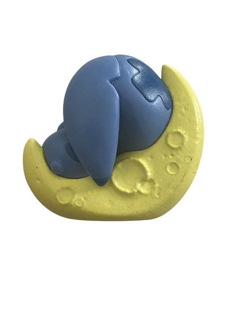 K&ouml;nnte beinhalten: Eine blaue Cartoonfigur liegt auf einem gelben Halbmond. Die Figur hat gro&szlig;e Ohren und einen runden K&ouml;rper. Der Mond hat eine strukturierte Oberfl&auml;che mit mehreren Kratern.