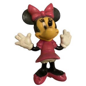 May include: A vintage Minnie Mouse figurine. The figure has a yellow face, black ears, and a pink bow. She wears a pink dress and black shoes. Her white gloved hands are raised. A classic Disney collectible.