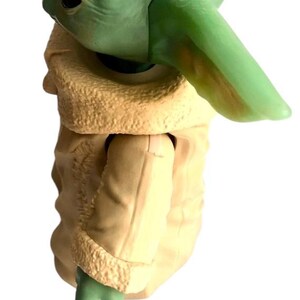 May include: A green and beige plastic toy figure of a character from the Star Wars franchise. The figure is wearing a brown coat with a fur trim.