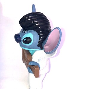 May include: A blue and white Stitch figurine wearing a white tuxedo and playing a brown guitar. The figurine has black hair styled like Elvis Presley.