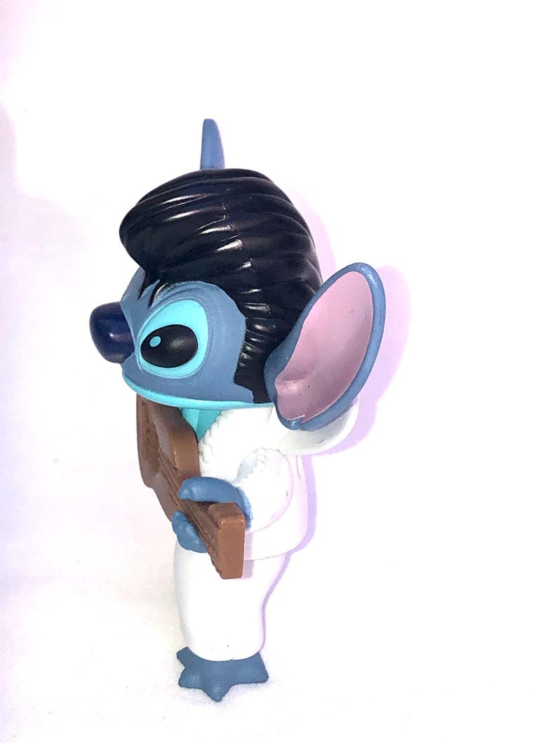 May include: A blue and white Stitch figurine wearing a white tuxedo and playing a brown guitar. The figurine has black hair styled like Elvis Presley.