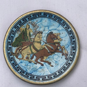 Puede incluir: Un pequeño plato redondo de cerámica azul y dorado con un borde de clave griega. El plato presenta una imagen pintada de una figura montando un carro tirado por cuatro caballos.
