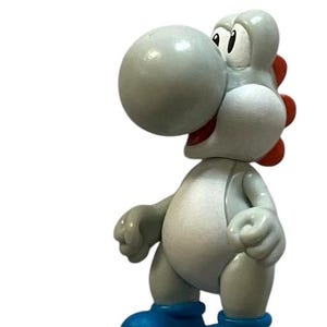 Pode incluir: Uma figura de Yoshi cinza e branca com uma cauda vermelha pontiaguda e sapatos azuis. A figura está em pé sobre um fundo branco.