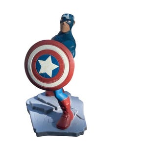 May include: A Captain America figurine in a dynamic pose, holding his iconic shield. The figure is predominantly blue, red, and white, standing on a gray base. The shield features a white star in a blue circle.