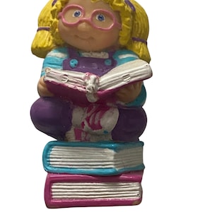 Könnte beinhalten: Figur einer Mädchenfigur mit blonden Zöpfen, rosa Brille, lila Latzhose und blau-weiß gestreiftem Oberteil. Sie liest ein Buch mit rosa Einband, das auf einem Stapel Bücher liegt.