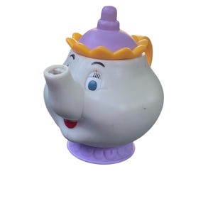 Puede incluir: Una tetera blanca de estilo caricatura con una cara sonriente, ojos azules y boca roja. La tapa es morada con un borde amarillo. La tetera se asienta sobre una base morada y tiene un asa amarilla.