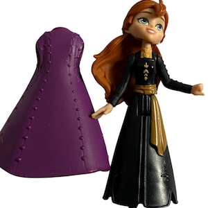 May include: A small plastic doll of a girl with long red hair wearing a black dress with gold accents and a purple dress. The doll is holding a purple dress in her left hand.