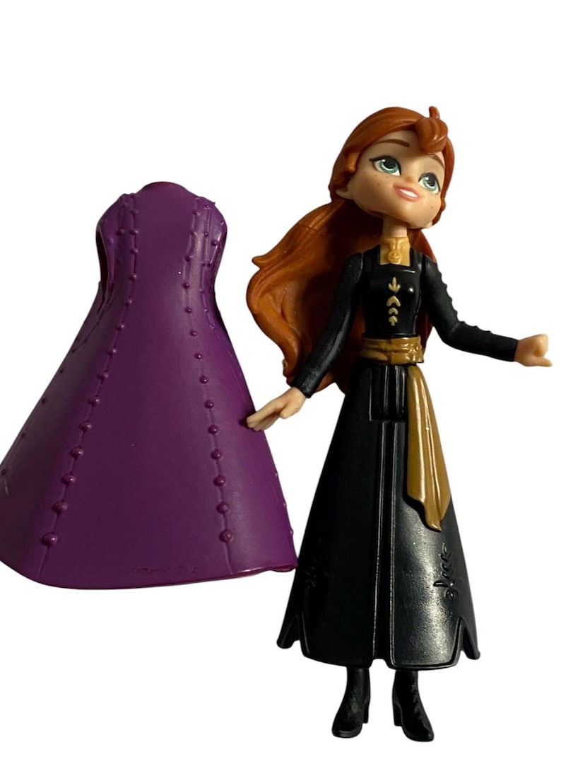 May include: A small plastic doll of a girl with long red hair wearing a black dress with gold accents and a purple dress. The doll is holding a purple dress in her left hand.