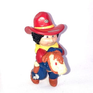 Puede incluir: Una figura de vaquero rojo, blanco y azul montando un caballo marrón y blanco. El vaquero lleva un sombrero de vaquero rojo con una banda amarilla y una camisa azul con un chaleco amarillo.